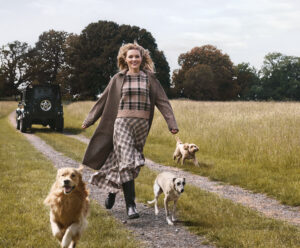 A blonde woman running through a field with a Labrador, a Whippet and a Cockapoo